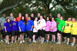 A diverse group of adults in colorful t-shirts posing together under a large leafy tree.