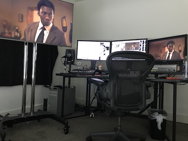 Video editing setup with three monitors on desks and a large screen displaying a man in a suit.