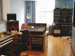 A music studio featuring a mixing console, rack equipment, a keyboard, and an electric guitar on the wall.