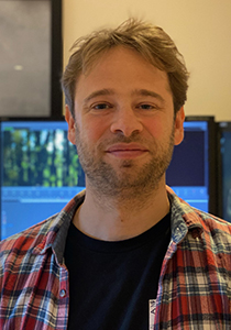 A man with light brown hair and a beard wearing a red and blue plaid shirt over a black t-shirt.
