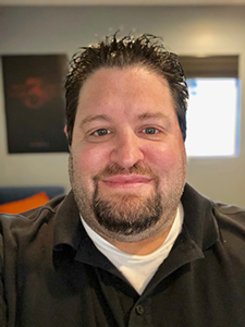 A smiling man with dark hair and a goatee wearing a black polo shirt over a white undershirt.
