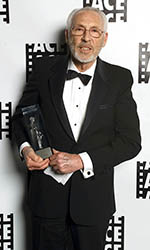 An older man with glasses and a beard wearing a black tuxedo and bowtie, holding a glass award.