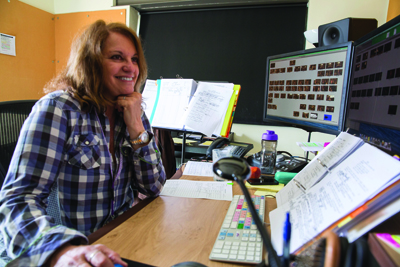 A smiling woman in a plaid shirt sits at a desk with computer monitors and sheet music.