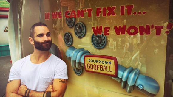 A man with a beard and white t-shirt stands before a sign with text and a cartoonish hand graphic.