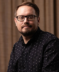 A man with short brown hair, glasses, and a goatee wearing a dark patterned button-down shirt.