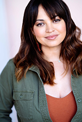 A woman with wavy dark hair and bangs smiles at the camera, wearing a green button-down shirt.