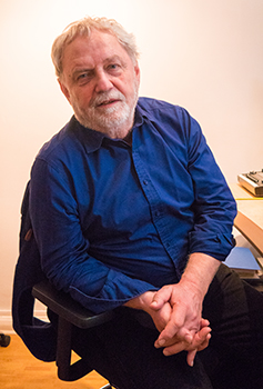 An older man with white hair and a beard wearing a blue button-down shirt sits in a chair.