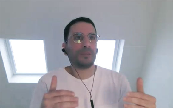 A man with glasses and wired earphones gestures while speaking in a video call.