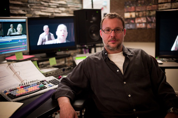 A man with glasses sits at a desk with multiple computer monitors and a video editing control surface.