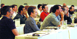 Several men sit at long tables in a brightly lit room, looking toward the front of a meeting.