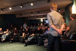 A man in a grey shirt stands with his back turned, facing a seated audience in a dimly lit room.