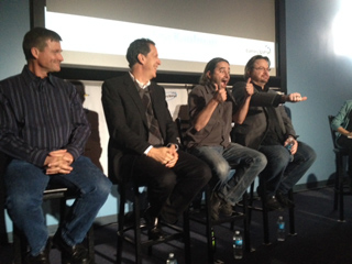 Four men sit on stools on a stage in front of a large projection screen.