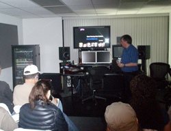A man in a blue shirt stands near a large monitor displaying graphics in a room with seated audience members.