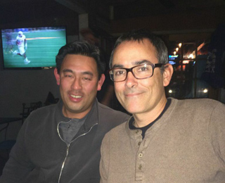 Two men smiling for a photo in a dark room with a television playing football in the background.