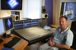 A man sits in a recording studio beside a large professional mixing console and multiple computer monitors.