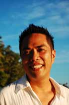 A smiling man with dark hair wearing a white collared shirt outdoors under a blue sky.