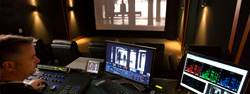 A man sits at a desk with multiple monitors and a control panel in a dark editing studio.
