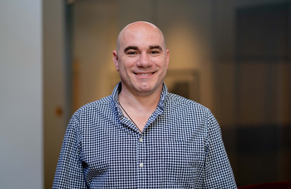 A smiling bald man wearing a blue and white checkered button-down shirt.