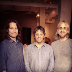 Three men with medium-length hair smiling at the camera in a dimly lit room.
