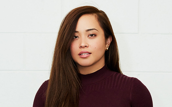 A woman with long brown hair and a maroon turtleneck looks toward the camera against a white tiled wall.