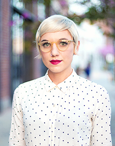 A woman with short blonde hair and yellow-rimmed glasses wearing a white polka dot shirt.