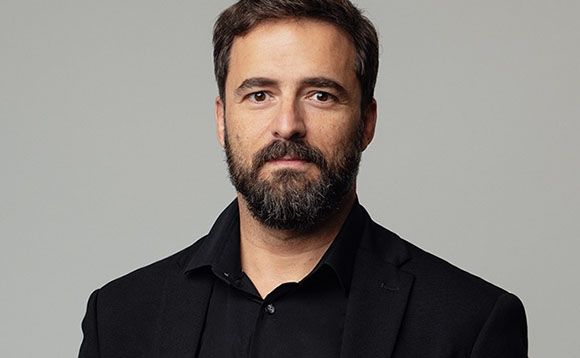 A man with dark hair and a full beard wearing a black blazer and shirt against a gray background.