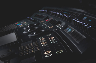 An angled view of a dark professional audio mixing console with illuminated buttons, sliders, and knobs.