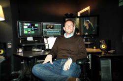 A man sits in a dark studio surrounded by computer monitors and professional audio equipment.