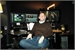 A person in a brown sweater and blue jeans sits in front of several computer monitors in a dark room.