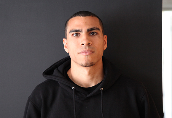 A man with short dark hair and a neutral expression wearing a black hoodie against a dark background.