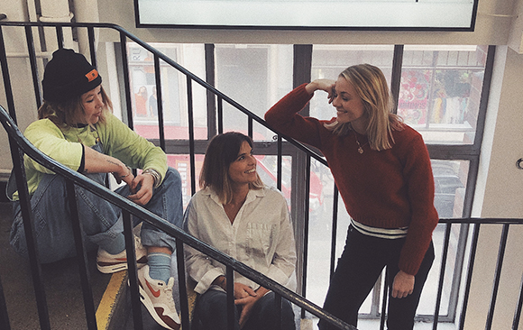 Three women smiling and talking on an indoor staircase near a large window.