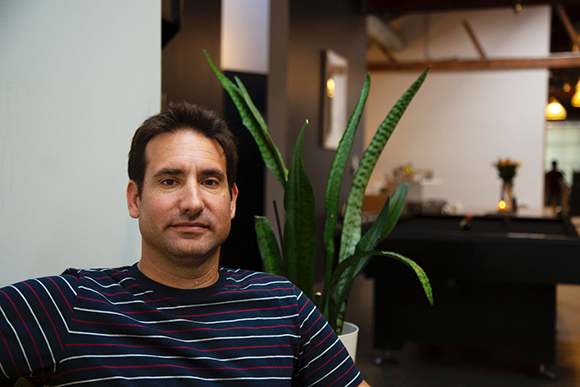 A man in a navy and red striped t-shirt sits in a room with a large snake plant behind him.