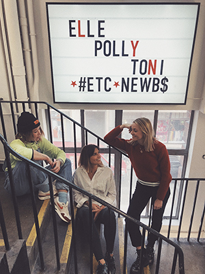 Three women pose on a staircase beneath a large light box sign with text.