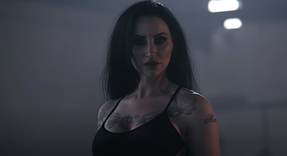 A woman with long dark hair and chest tattoos looks forward in a dimly lit setting.