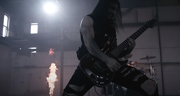A person in dark clothing plays a black electric guitar in a smoky room with flames behind them.
