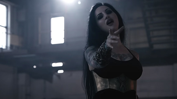 A woman with long dark hair and dark makeup reaches her hand toward the camera in a dimly lit room.