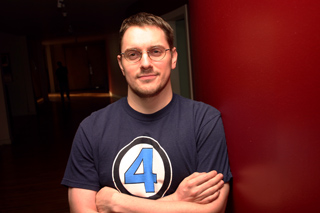 A man with glasses and a beard wearing a blue t-shirt with a large number 4 logo, standing against a red wall.