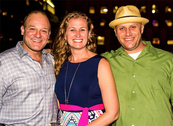 A man in a plaid shirt, a woman in a blue dress, and a man in a green shirt and straw hat smiling.
