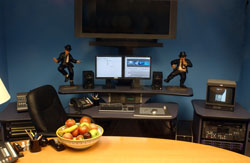 A desk with dual computer monitors, speakers, two action figurines, and a small television in a blue room.
