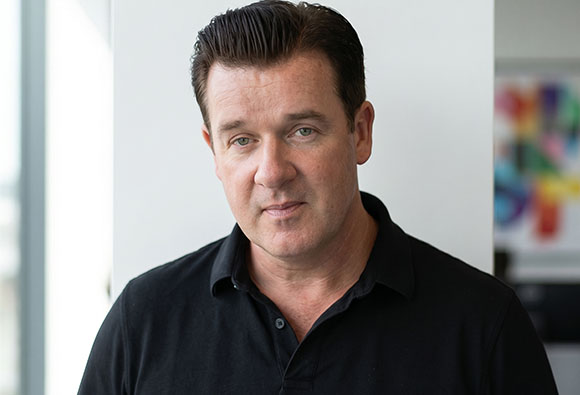A man with short dark hair wearing a black polo shirt looking forward.