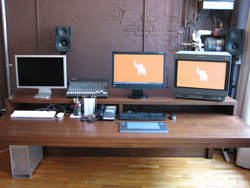 A wooden desk with three monitors, a mixing board, speakers, and a computer tower.