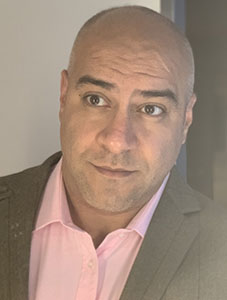 A bald man wearing a pink collared shirt and a grey blazer looks off-camera with a slight smile.