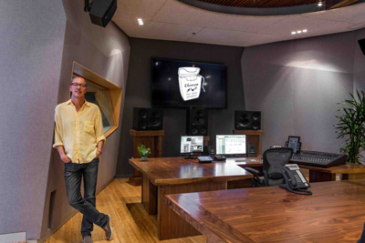 A man in a yellow shirt leans against a wall in a recording studio with large wooden desks and monitors.