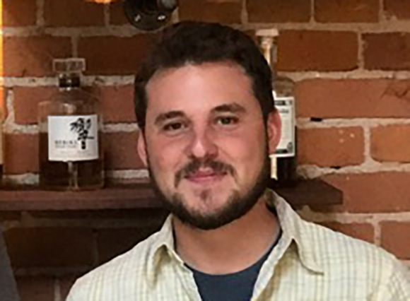 A man with dark hair and a beard smiling in a light-colored button-down shirt.