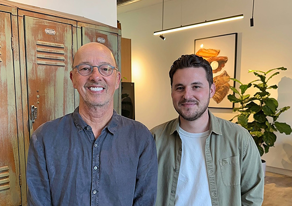 A bald man with glasses and a younger man with dark hair smiling in front of lockers and a plant.