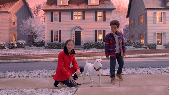 A girl in a red coat and a boy in a plaid jacket stand by a white Bull Terrier on a snowy sidewalk.