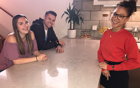 A woman in red, a man in a black hoodie, and a woman in a purple top smiling behind a kitchen counter.