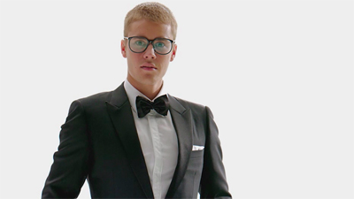 A young man wearing a black tuxedo, white shirt, black bowtie, and glasses against a white background.