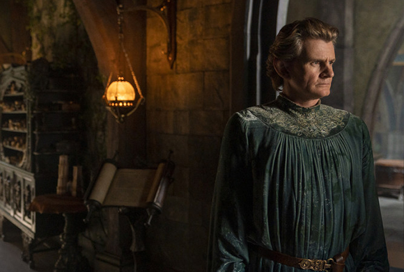 A man with wavy hair wearing a green pleated tunic stands in a stone room next to a hanging lantern.