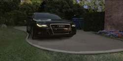 A black Audi sedan driving along a curved residential driveway at dusk.